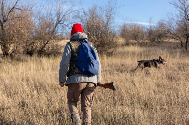 Olgun bir avcı elinde pompalı tüfekle tarlada yürüyor.