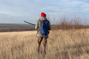 Olgun bir avcı elinde pompalı tüfekle tarlada yürüyor.