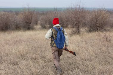 Olgun bir avcı elinde pompalı tüfekle tarlada yürüyor.