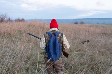 Olgun bir avcı elinde pompalı tüfekle tarlada yürüyor.
