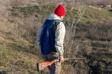 Olgun bir avcı elinde pompalı tüfekle tarlada yürüyor.