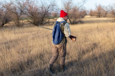 Olgun bir avcı elinde pompalı tüfekle tarlada yürüyor.