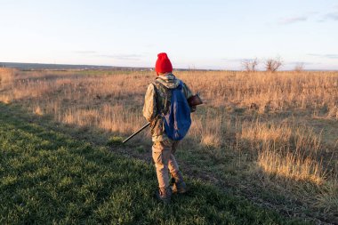 Olgun bir avcı elinde pompalı tüfekle tarlada yürüyor.