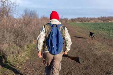 Olgun bir avcı elinde pompalı tüfekle tarlada yürüyor.