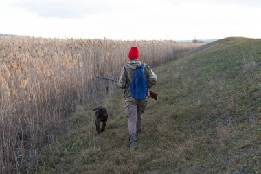 Olgun bir avcı elinde pompalı tüfekle tarlada yürüyor.