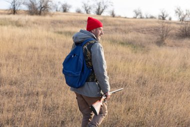 Olgun bir avcı elinde pompalı tüfekle tarlada yürüyor.