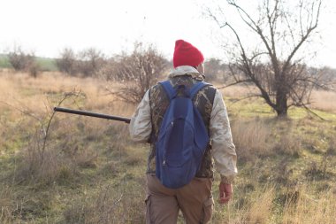 Olgun bir avcı elinde pompalı tüfekle tarlada yürüyor.