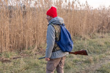 Olgun bir avcı elinde pompalı tüfekle tarlada yürüyor.