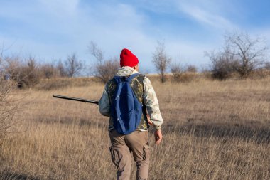Olgun bir avcı elinde pompalı tüfekle tarlada yürüyor.