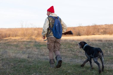 Olgun bir avcı elinde pompalı tüfekle tarlada yürüyor.
