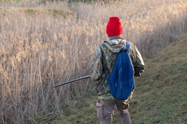 Olgun bir avcı elinde pompalı tüfekle tarlada yürüyor.