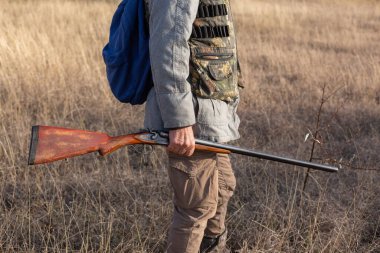Olgun bir avcı elinde pompalı tüfekle tarlada yürüyor.