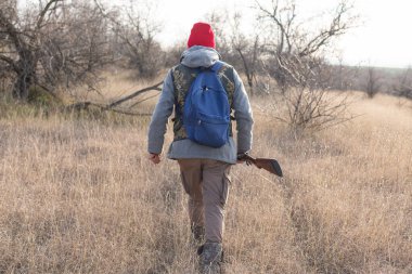 Olgun bir avcı elinde pompalı tüfekle tarlada yürüyor.