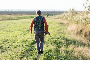 Olgun bir avcı elinde pompalı tüfekle tarlada yürüyor.
