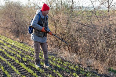 Olgun bir avcı elinde pompalı tüfekle tarlada yürüyor.