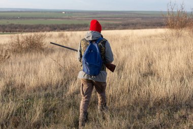 Olgun bir avcı elinde pompalı tüfekle tarlada yürüyor.