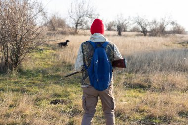 Olgun bir avcı elinde pompalı tüfekle tarlada yürüyor.