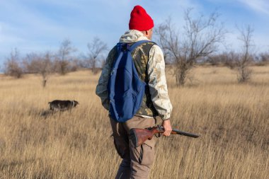 Olgun bir avcı elinde pompalı tüfekle tarlada yürüyor.