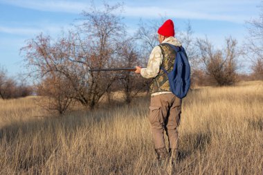 Olgun bir avcı elinde pompalı tüfekle tarlada yürüyor.