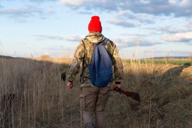 Olgun bir avcı elinde pompalı tüfekle tarlada yürüyor.