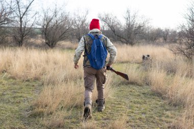 Olgun bir avcı elinde pompalı tüfekle tarlada yürüyor.