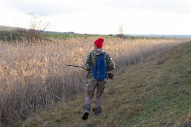 Olgun bir avcı elinde pompalı tüfekle tarlada yürüyor.