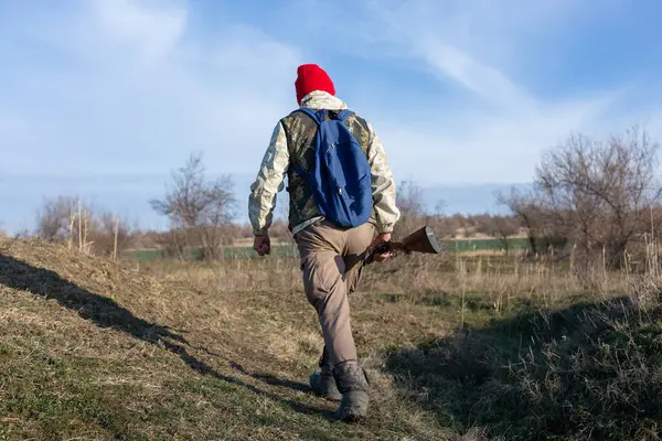 Olgun bir avcı elinde pompalı tüfekle tarlada yürüyor.