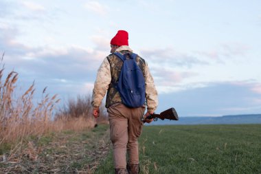 Olgun bir avcı elinde pompalı tüfekle tarlada yürüyor.