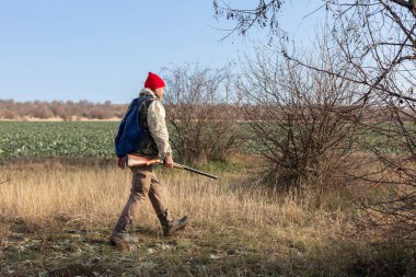 Olgun bir avcı elinde pompalı tüfekle tarlada yürüyor.