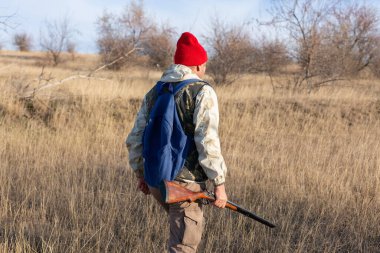 Olgun bir avcı elinde pompalı tüfekle tarlada yürüyor.