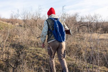 Olgun bir avcı elinde pompalı tüfekle tarlada yürüyor.