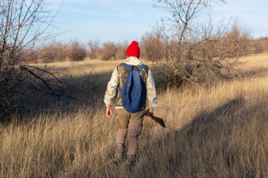 Olgun bir avcı elinde pompalı tüfekle tarlada yürüyor.