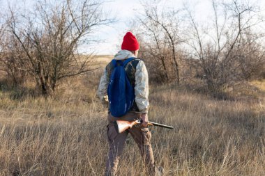 Olgun bir avcı elinde pompalı tüfekle tarlada yürüyor.
