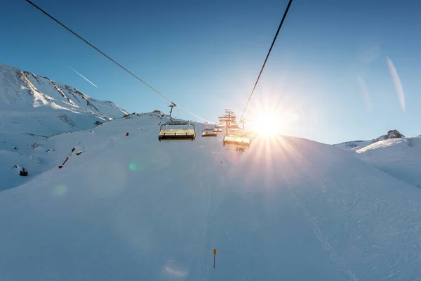 阳光明媚的夜晚 滑雪车在高山山地冬季度假胜地空旷的缆车上 滑雪椅拉索的方式与人们喜欢滑雪和滑雪板 夕阳西下的天空在背景上闪烁着光芒 — 图库照片
