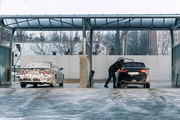 Men driver at car wash with high pressure water equipment pump at self-service outdoor cold snow frosty winter day. Vehicle covered with foam shampoo chemical detergents during carwash self service.