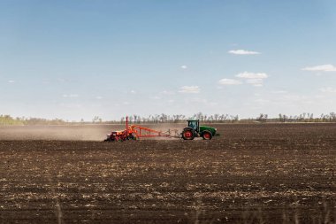 Green big modern tractor covered with earth dust seeding agricultural field at farm on bright sunny day. Farmer cultivating and make soil tillage before seeding plants crops. Countryside rural scene.