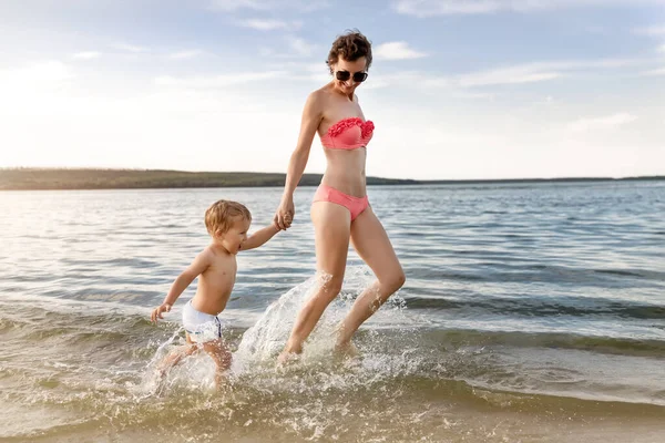 Young adult attractive slim sporty mother enjoy having fun running water by lake or sea sand breach with cute little baby boy against blue sky on summer day. Summertime family vacation concept.