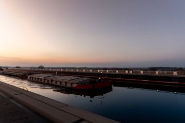 Manzara, Alman iç kanalı Magdeburg Köprüsü 'nden geçen büyük kargo yük römorkörü mavnası mavnası gemisinin sıcak günbatımı gökyüzüne karşı çekildiğini gösteriyor. Su taşımacılığı lojistik nakliye endüstrisi Avrupa.