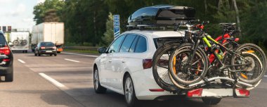 Back view modern white familywagon car with mounted roof box trunk and bike tail carrier driving european highway road against blue sky summer day. Lifestyle travel adventure trip journey concept.