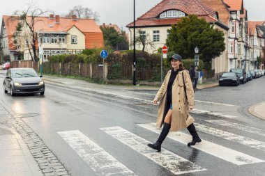 Genç, modaya uygun, şık, güzel, beyaz, mutlu, gülümseyen kadın yağmurlu bir günde Avrupa 'da yaya geçidinde yürümeyi seviyor. Kadın portresi, şehir caddesinde kot pantolon giyer, trençkot giyer..