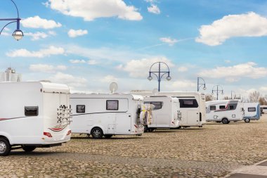 Many white modern campervan recreational motor home vehicles parked in row at camper park site Magdeburg city against Elbe river bridge. Motorhome campground stataion travel destination. RV lifestyle.