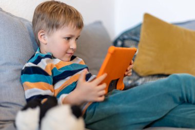 Sevimli, sevimli sarışın çocuğun portresi evde kanepede oturmaktan, internette sörf yapmaktan ya da tablet kullanarak TV dizisi filmleri izlemekten zevk alıyor. Çocuklar akıllı teknoloji kullanımı konsepti. Çocuk aleti bağımlılığı.