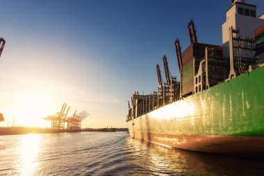 Manzaralı dev kargo konteynır gemisi Hamburg limanını liman vinçlerine karşı sıcak akşam gün batımına karşı dolduruyor. Küresel ticari taşımacılık kiralama ve lojistik geçmişi.