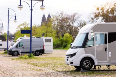 Many white modern campervan recreational motor home vehicles parked in row at camper park site Magdeburg city against Elbe river bridge. Motorhome campground stataion travel destination. RV lifestyle.