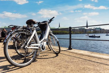 Scenic view two modern bicycles parked at embankment of Alster lake in Hamburg street old city center background summer day panorama view. Healthy travel lifestyle concept. Urban rental transport. clipart