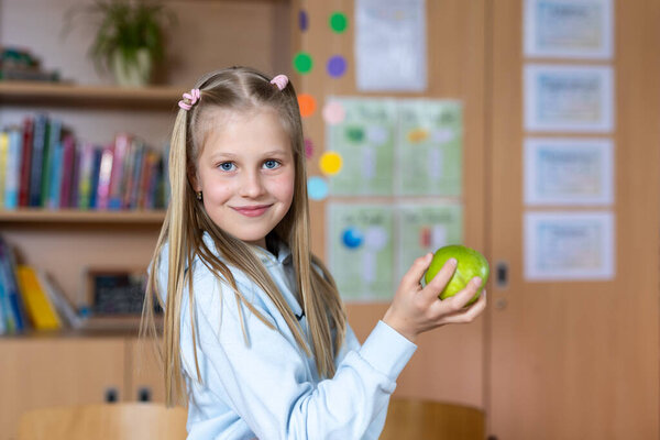 Portrait cute blond confident happy caucasian little blond little kid girl hold hand eating apple in classroom at lunch time pause. Schoolchild in class. Healthy snack food. Back to school concept.