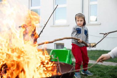 Sevimli küçük sevimli oğlan bahçesinde büyük bir ateş olan büyük tahta çubuğu ateş çukuruna koydu. Çocuk şenlik ateşiyle tehlikeli bir oyun oynuyor. Yangın kazası riski ve sağlık damgae konsepti.