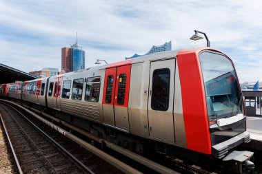 Hamburg Red Unmanned U-bahn Metro treni Hamburger Limanı Elbphilharmonie 'nin arka planındaki Yükseltilmiş Pist' teki Baumwall istasyonunda mavi gökyüzü. Hanseatik şehri ulaşım manzaralı.