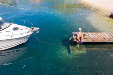 Şirin küçük kız, ahşap iskelede oturuyor. Manzaralı turkuaz deniz suyuyla dolu yatın yanında. Mutlu çocuk ağda balık tutarken eğlen yaz tatilinde doğayı keşfet..