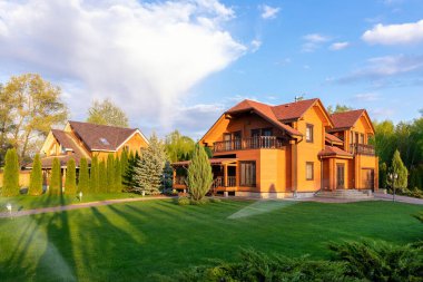 Modern wooden eco house villa facade luxury big house. Timber cottage with with green lawn water sprinkler, paved footpath and blue sky background. Landscaping design, garden watering and maintenance.